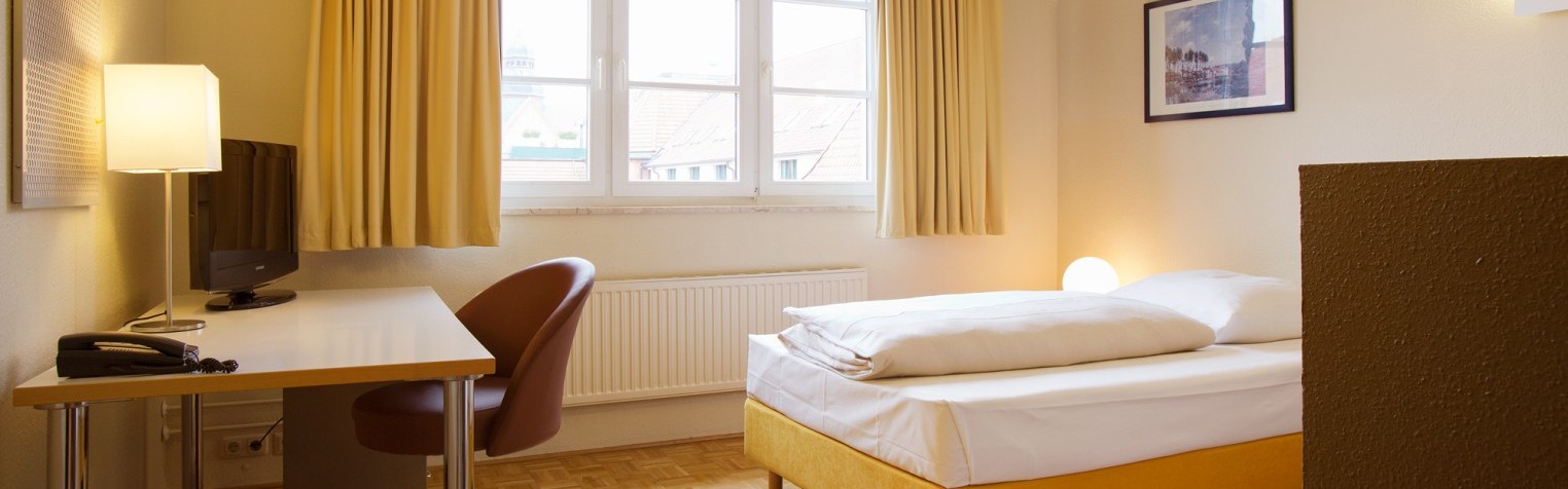 Einladend eingerichtetes Einzelzimmer mit einem Schreibtisch, Sessel, Fernseher und gelben Bett am Fenster