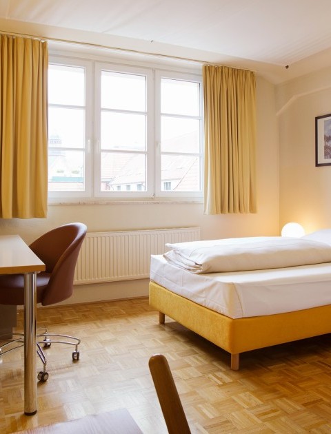 Einladend eingerichtetes Einzelzimmer mit einem Schreibtisch, Sessel, Fernseher und gelben Bett am Fenster