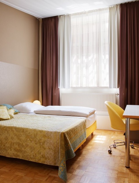 Ein helles möbliertes Zimmer Hannover mit einem Doppelbett, Schreibtisch, Fernseher und Fenster mit Vorhängen