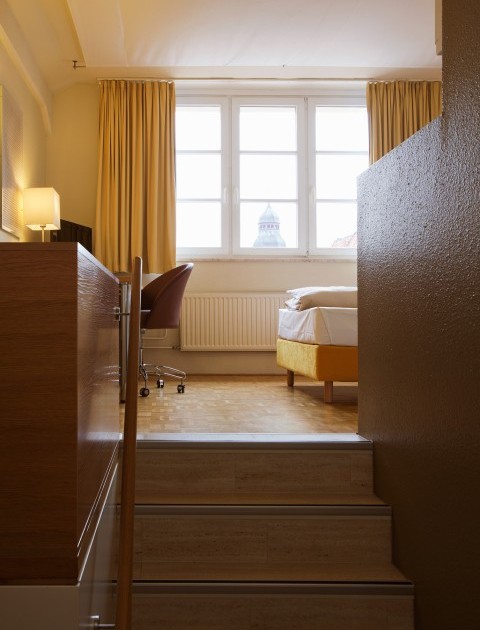 Helles Einzelzimmer in Hannover mit Schreibtisch und gelbem Bett, erreichbar über eine kleine Treppe im Raum