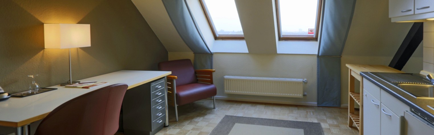 Ein helles Zimmer mit Dachfenstern, einem Schreibtisch, Sessel und einer integrierten Küchenzeile mit Spüle