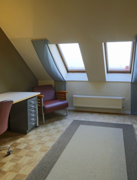 Ein helles Zimmer mit Dachfenstern, einem Schreibtisch, Sessel und einer integrierten Küchenzeile mit Spüle